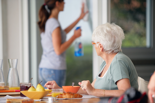 senior woman having breakfast, caregiver cleaning windows