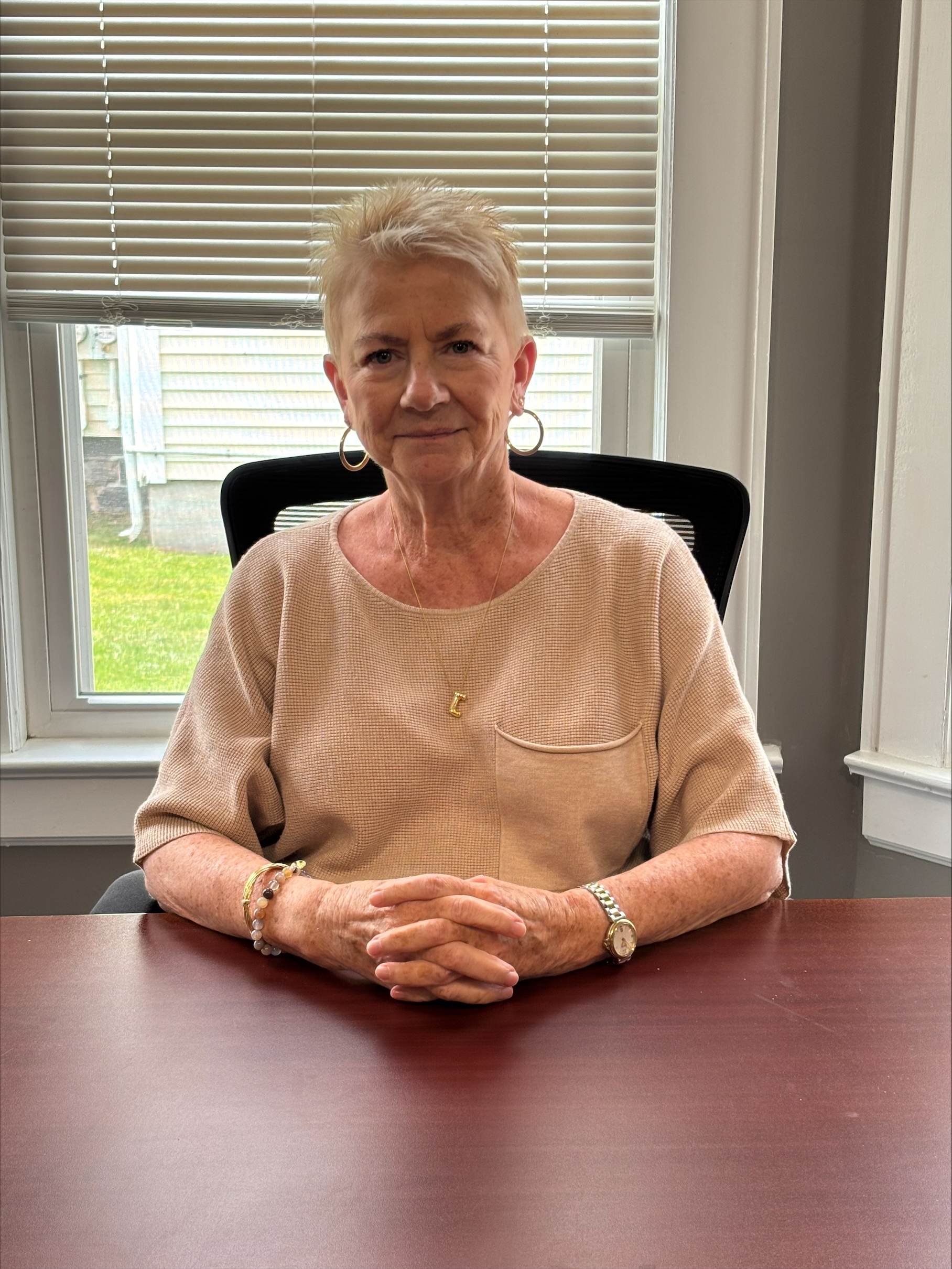 A headshot of Joy Thompson sitting at a table. Her hands are crossed in front of her, and there is a window behind her.