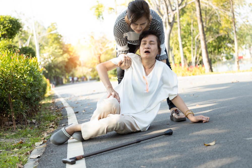 A concerned woman helps an elderly woman who has collapsed on a paved park path, possibly due to heat exhaustion; a walking cane lies on the ground nearby.