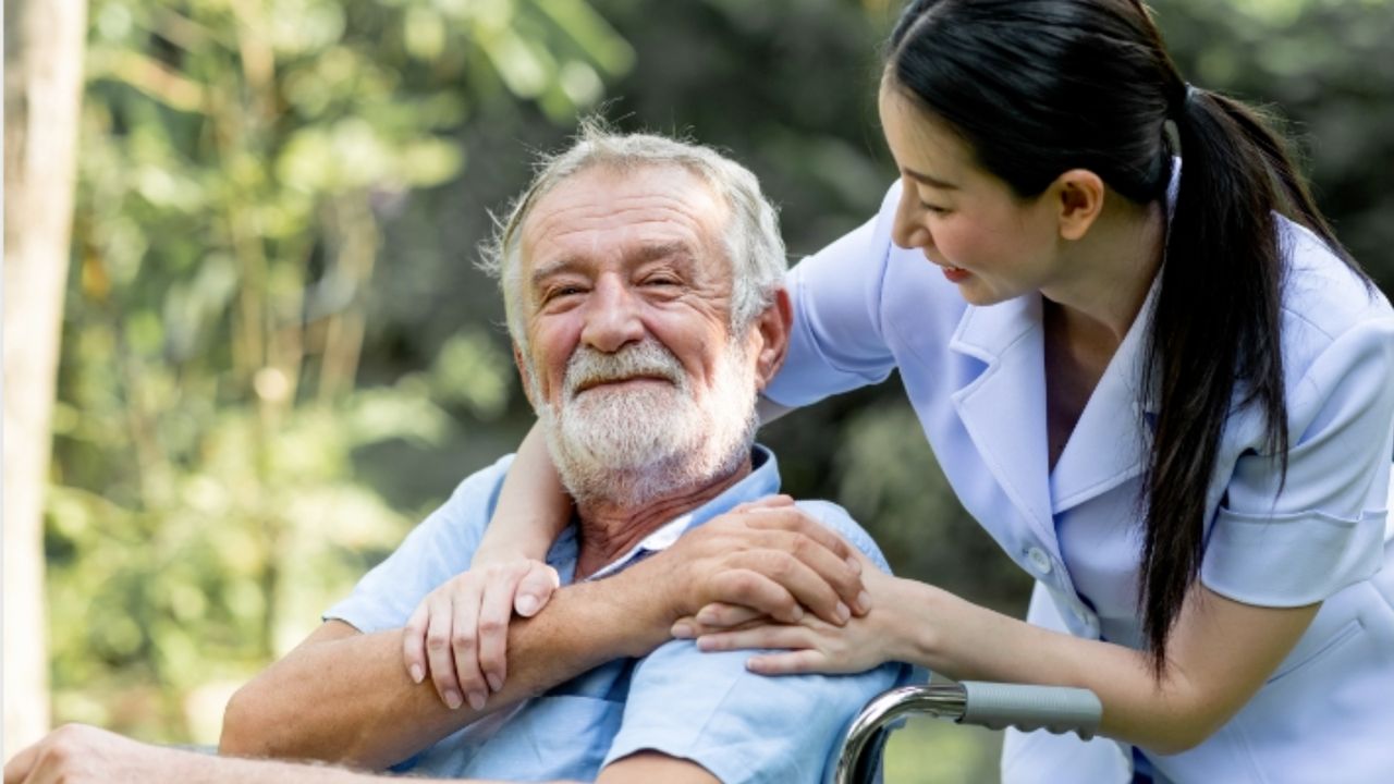 A smiling elderly man in a wheelchair is being comforted by a nurse from the home health care service, who is leaning over him and placing her hands on his shoulders.