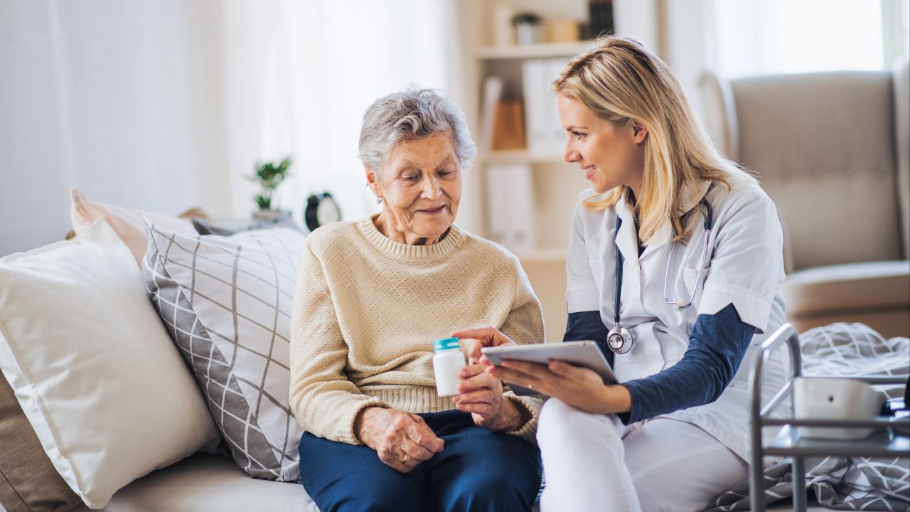 A nurse in uniform sits on a couch next to an elderly woman, holding a container of medication and showing something on a tablet. The elderly woman, benefiting from elder home care services, is seated and observing the tablet.