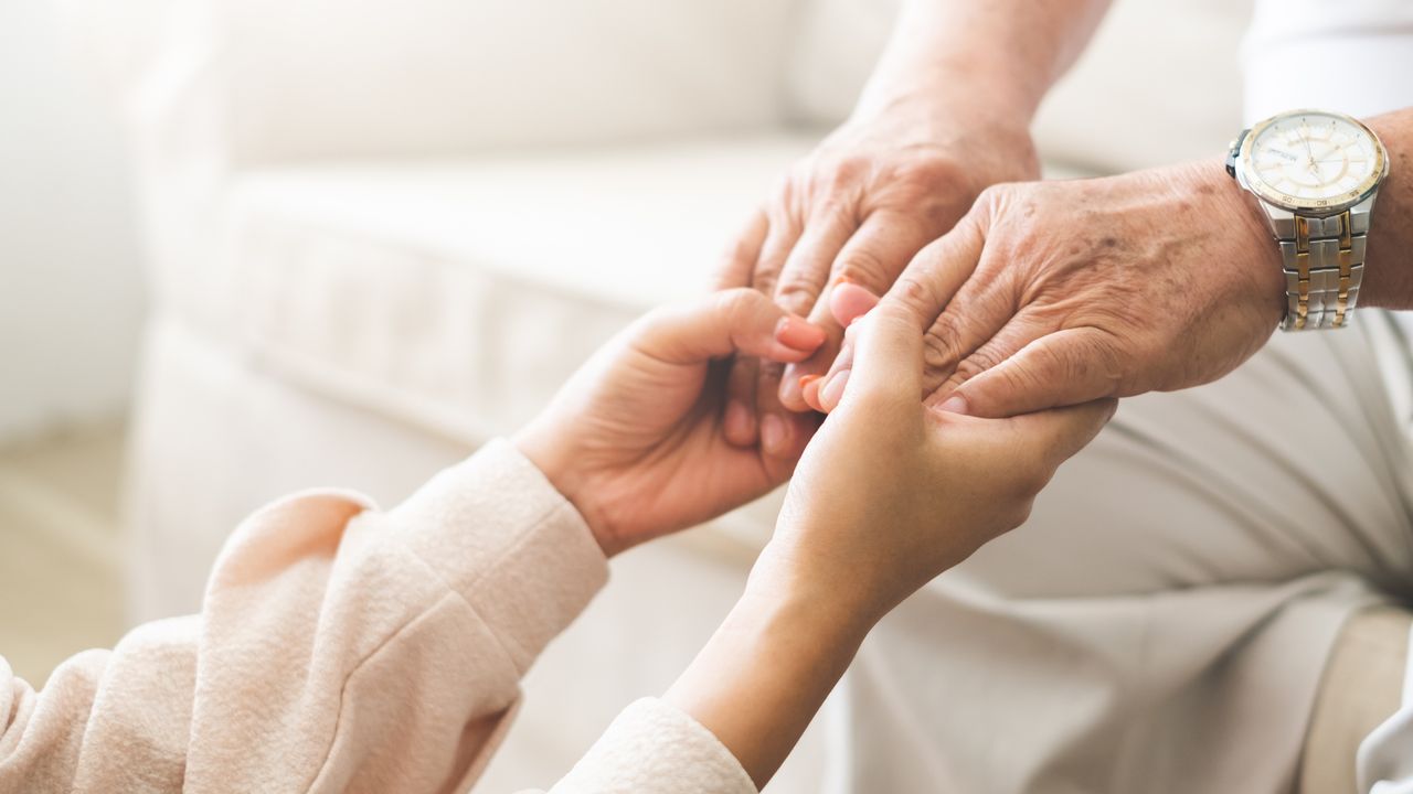 A young person holds the hands of an older individual, both sitting on a light-colored sofa, embodying the essence of personal care services. The older person wears a watch.