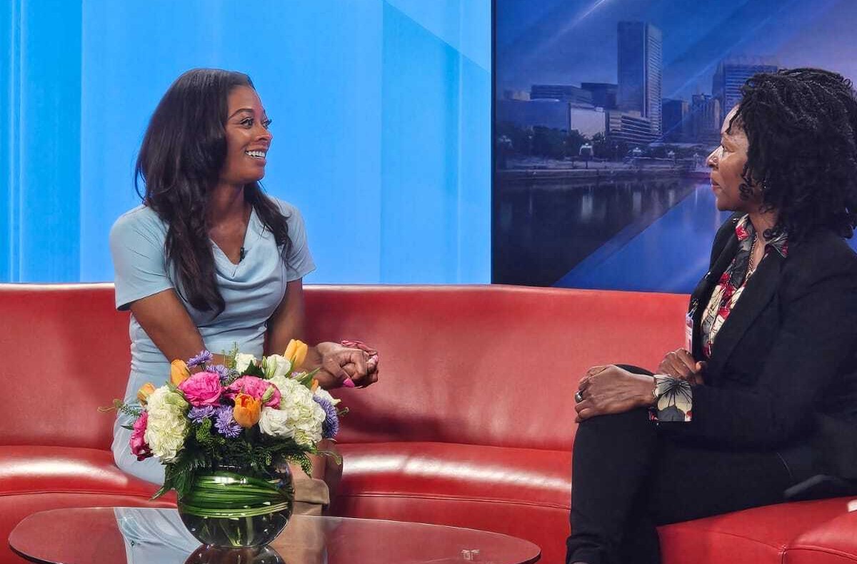 Two women are seated on a red couch in a studio, engaged in conversation about elder home care services. A vase with colorful flowers is placed on the table in front of them.