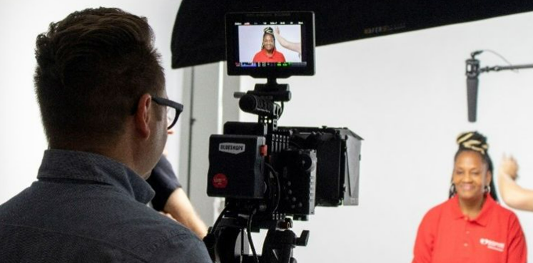 A person in a red shirt is being filmed in a studio setting focused on home care. A camera operator is adjusting the camera while viewing the subject on a monitor.
