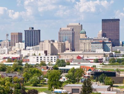 A city skyline featuring several mid to high-rise buildings under a partly cloudy sky, with a foreground of trees and low-rise buildings that house various caregiver services.
