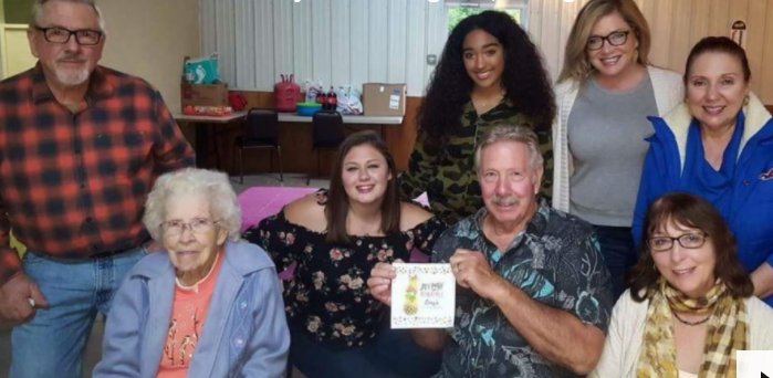 Group photo of seven adults and one elderly woman. The elderly woman, representing the spirit of home care, is seated in front next to a man holding a framed item. Four women and one man stand behind them, all smiling with the warmth of personal care services they've received.