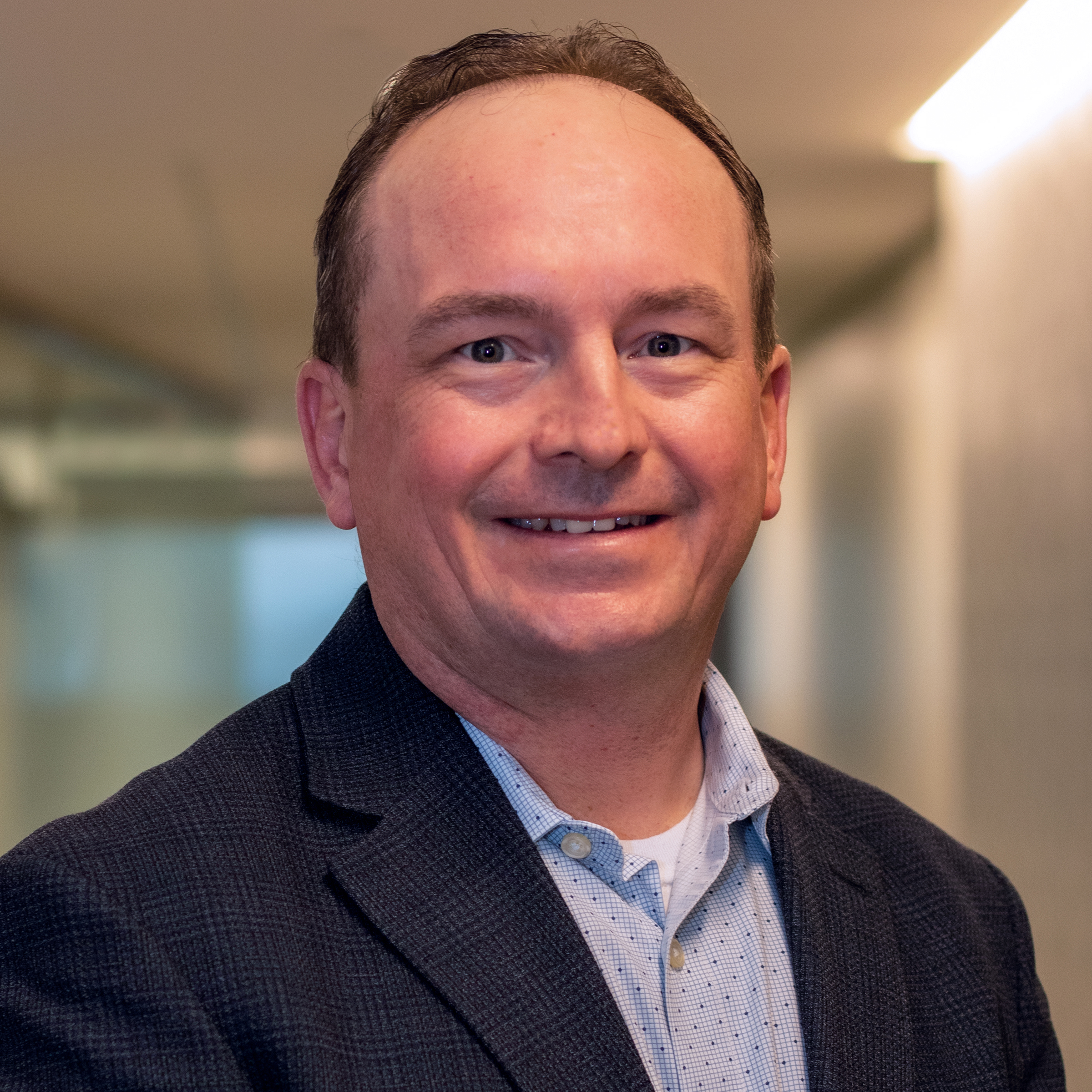 A man with short hair wearing a dark blazer and light blue shirt smiles at the camera in an indoor setting, where he provides home health care services to those in need.