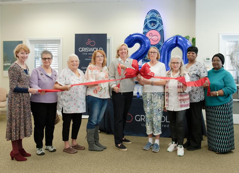 A group of women stands in a room, holding a red ribbon for a ceremonial ribbon-cutting event. Blue balloons with a "40" are in the background along with Griswold Home Care signage, celebrating four decades of exceptional caregiver services.