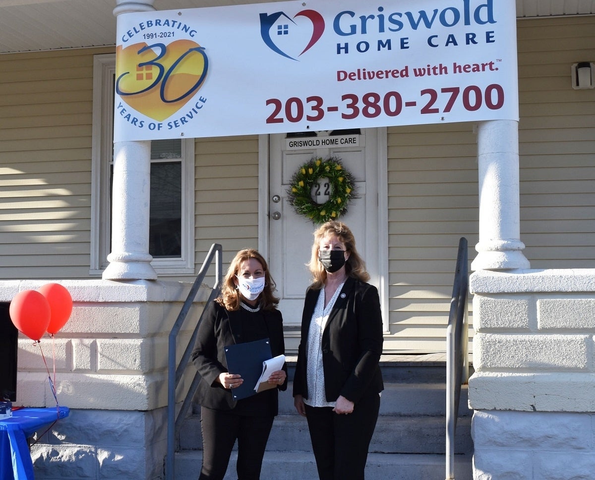 Two women wearing masks stand in front of a Griswold Home Care building. The banner above them reads, "Celebrating 30 Years of Service," highlighting their dedication to elder home care services and listing contact details including a phone number.