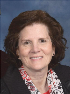A woman with short, wavy brown hair, wearing a black sweater and a patterned scarf, smiles at the camera against a blue background, embodying the warmth and compassion of our personal care services.