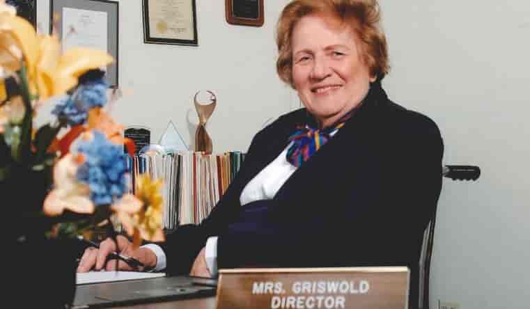 A woman wearing a suit is seated at a desk with a nameplate that reads "Mrs. Griswold Director." Surrounded by flowers and certificates, she exemplifies the dedication to home care and personal care services.